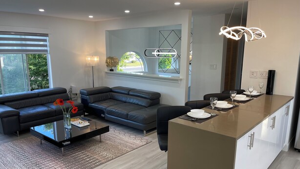 A modern living room with a grey sofa, a coffee table, and a dining area with a long table set for two. - Cozy , spacious and comfortable house. Pet friendly! And close to everything!!! (Chilliwack)
