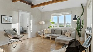 Ein geräumiges Wohnzimmer mit hohen Decken, einem grauen Sofa, einem Holztisch und einem großen Fenster mit Blick auf Grünflächen.