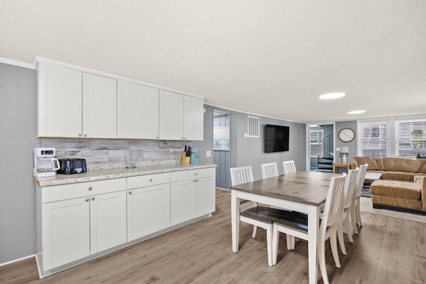 A modern kitchen with white cabinets, a dining area with a wooden table and chairs, and a living room with a beige sofa.