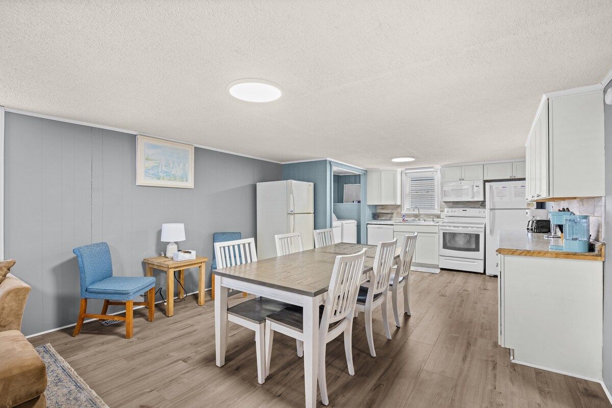 A modern kitchen with white appliances, a dining area with a wooden table and chairs, and a living room with a blue chair and a sofa.