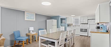 A modern kitchen with white appliances, a dining area with a wooden table and chairs, and a living room with a blue chair and a sofa.