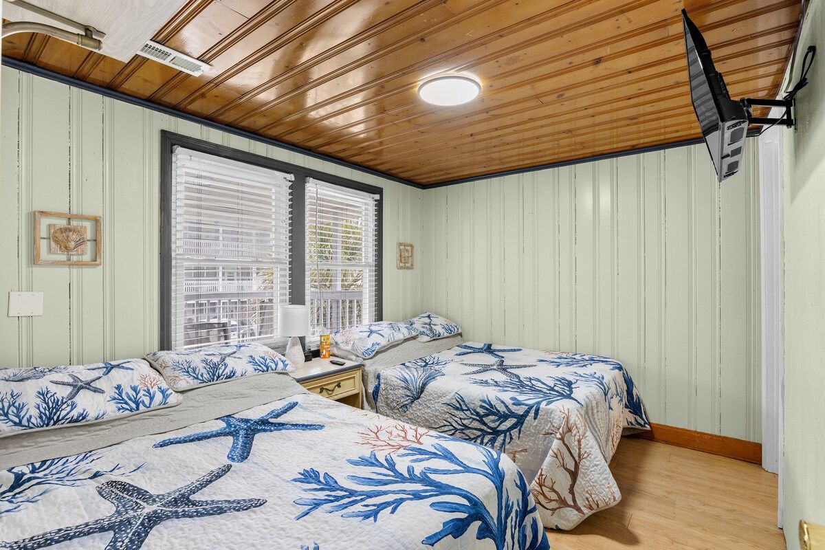 Ein Zimmer mit zwei Betten, einem Fenster mit Jalousien, einem Deckenventilator und einem an der Wand montierten Fernseher.