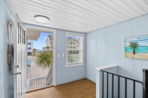 Ein Zimmer mit Holzboden, einer weißen Tür, einem Fenster mit Jalousien und einem Gemälde, das eine Strandansicht zeigt.