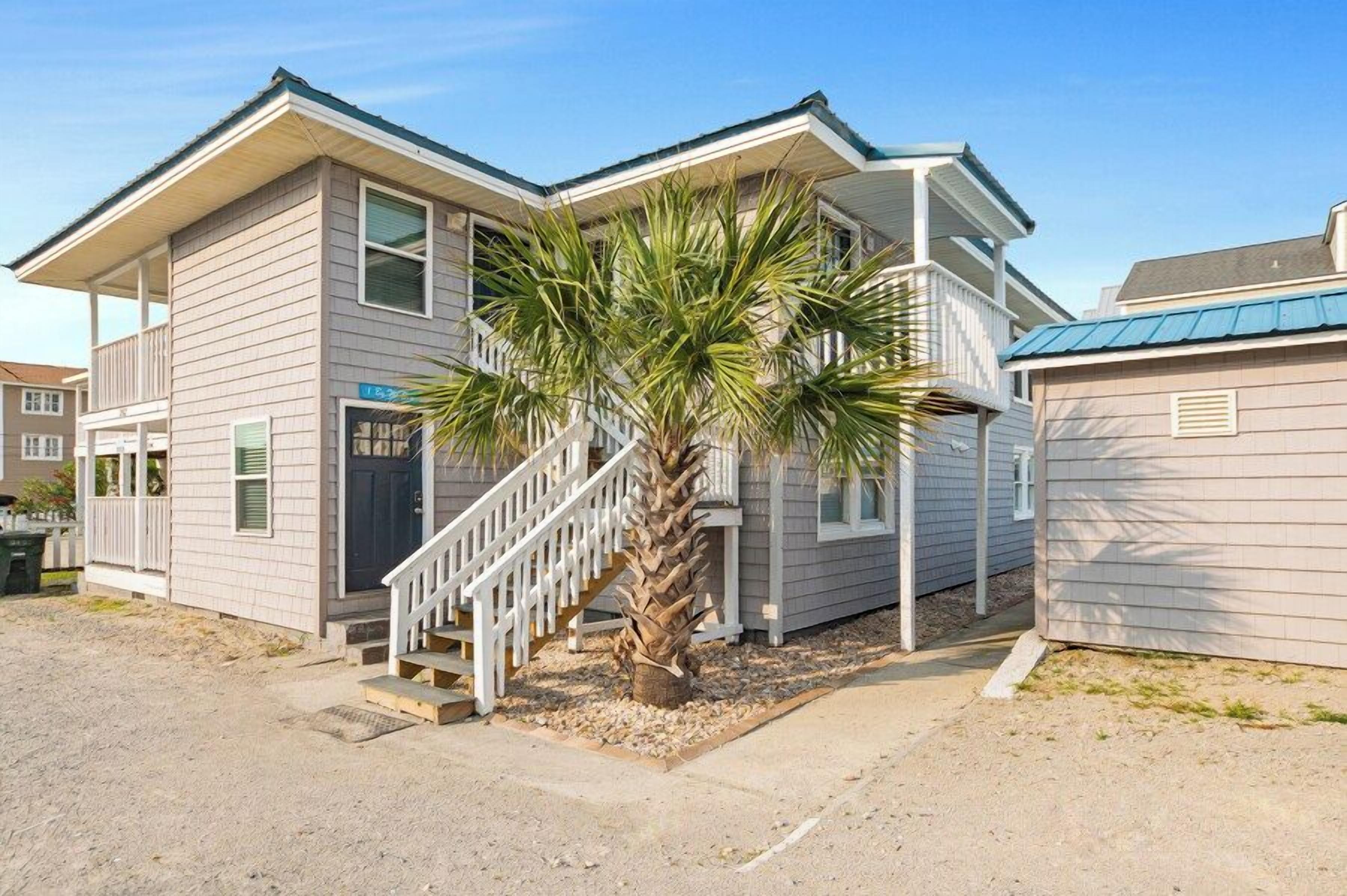 A two-story building with a staircase leading to the entrance, a palm tree in front, and a clear sky.