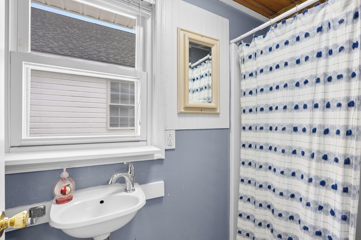 A bathroom with a white sink, a mirror, a window with blinds, and a shower curtain with a blue pattern.