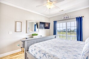 A bedroom with a bed, a ceiling fan, a TV, a window with curtains, and a view of the outside.