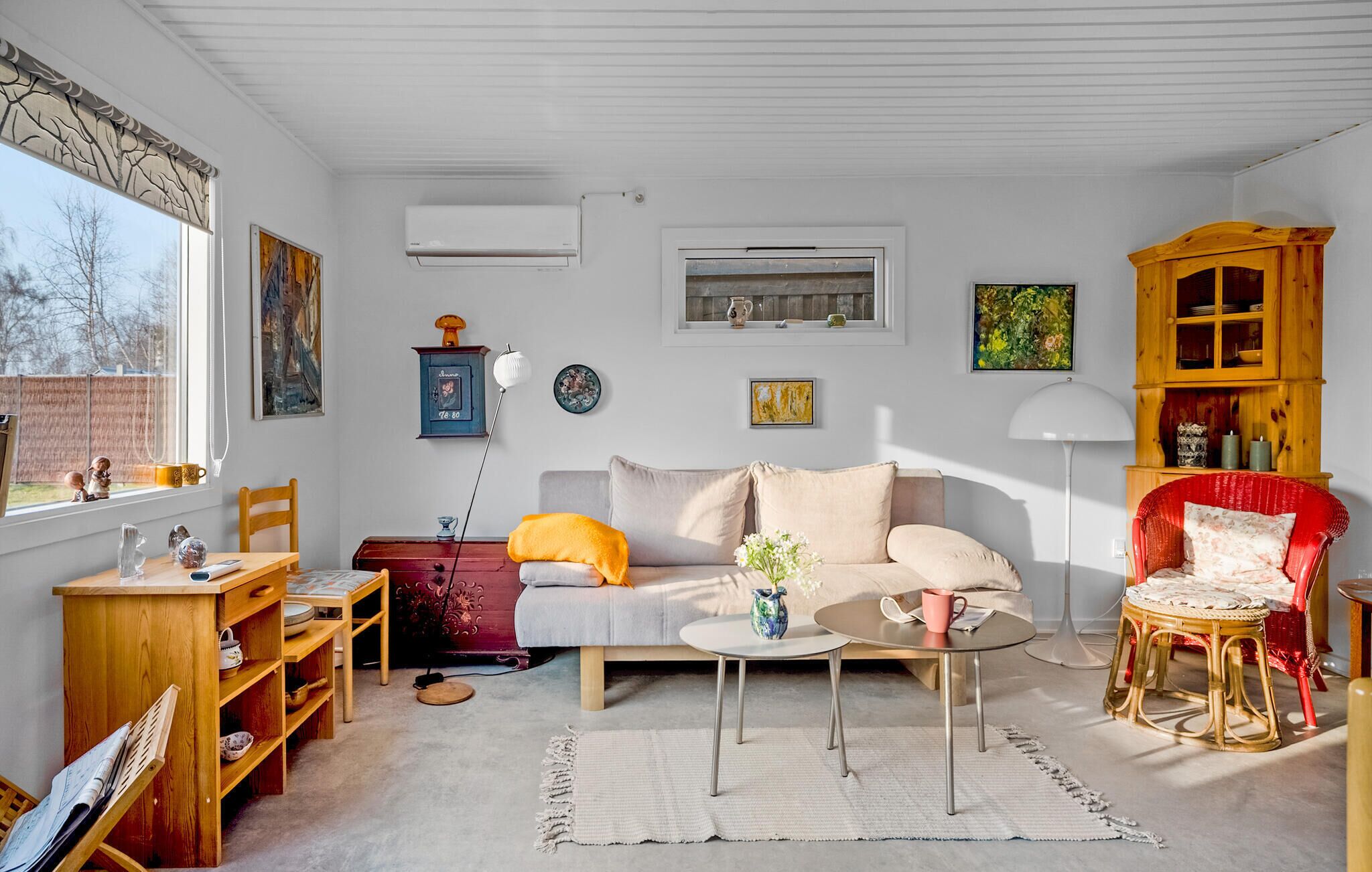 Un salon chaleureux avec un canapé, une table basse, un fauteuil rouge, un meuble en bois et une fenêtre donnant sur des arbres.