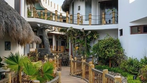 Un resort con una terraza de madera que conecta varias edificaciones, rodeado de exuberante vegetación y construcciones con techo de paja.