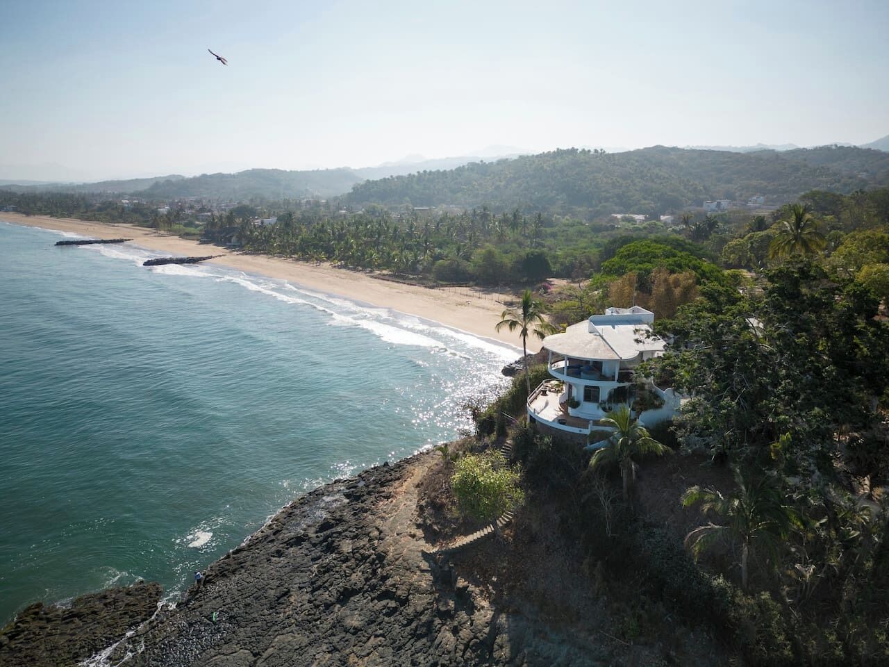 A coastal property with a beach view, a house with a balcony overlooking the sea, and a mountainous backdrop.