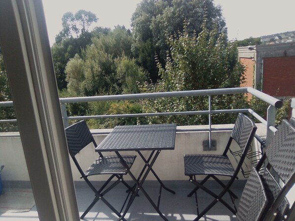 Un balcón con una mesa y cuatro sillas, con vistas a un jardín con árboles y un edificio al fondo.