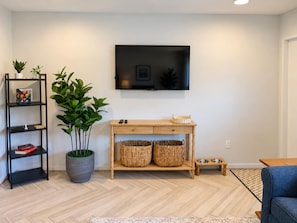 A living room with a flat-screen TV mounted on the wall, a wooden console table with baskets, a black shelving unit with books, and a potted plant.