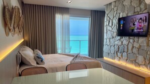 A modern hotel room with a large bed, a glass table, a stone accent wall, a TV displaying a show, and a view of the ocean. - RN Studio Premium 613 - Frente Mar (Maceió)
