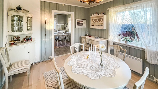 A dining area with a round table, white chairs, and a lace table runner. There is a cabinet with glass doors and a window with a view of greenery. - Cozy home in Gravberget (Gravberget)