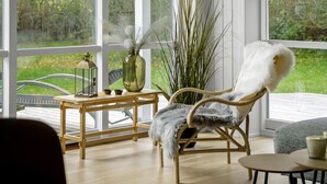 Ein gemütliches Zimmer mit einem großen Fenster, ein Korbstuhl mit einem flauschigen Kissen, ein Holztisch mit einer Laterne und eine Vase mit Trockenpflanzen.