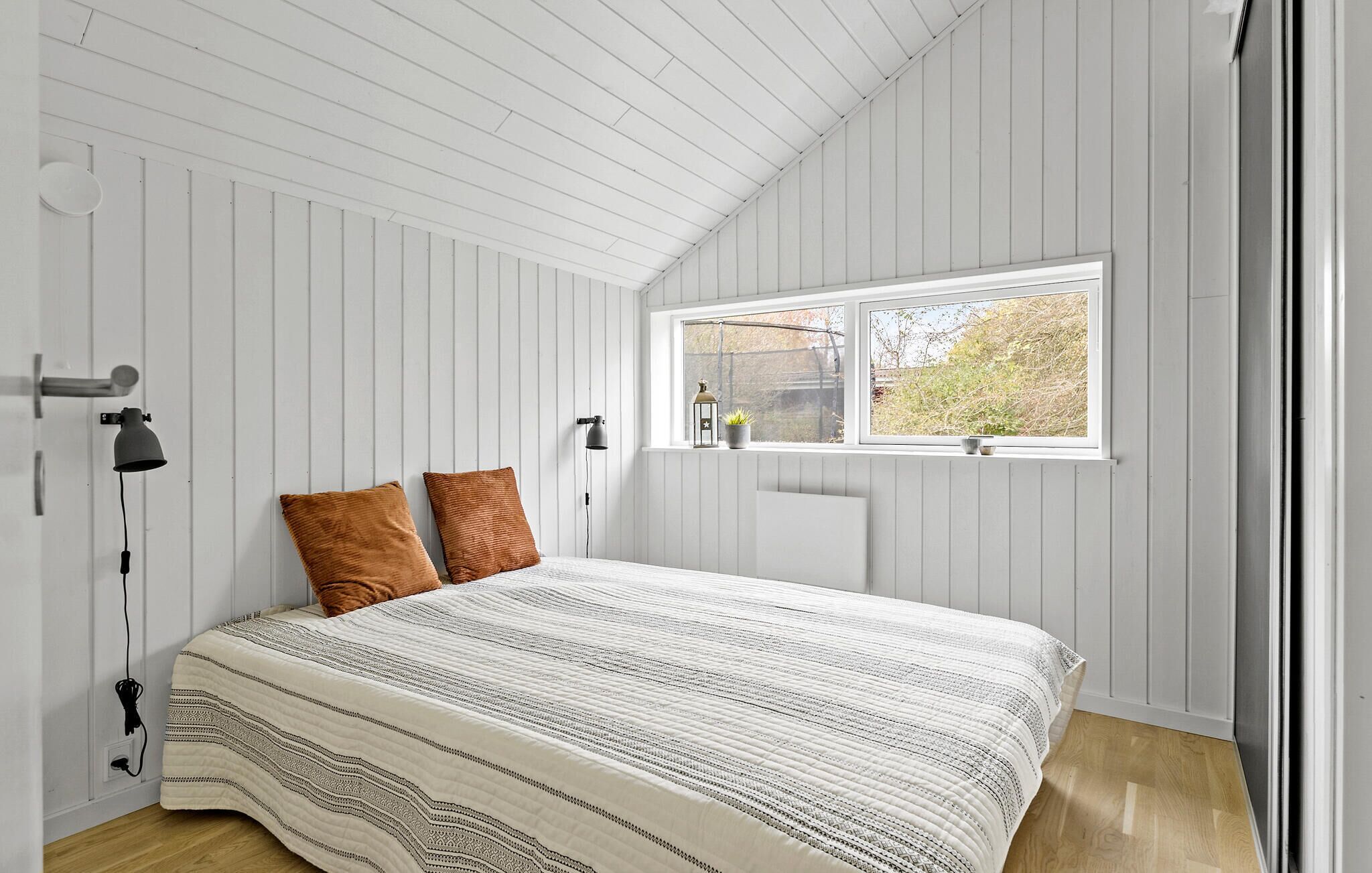 Ein Schlafzimmer mit einem Bett, zwei Kissen, einem Nachttisch, einer Lampe und einem Fenster mit Blick auf Bäume.