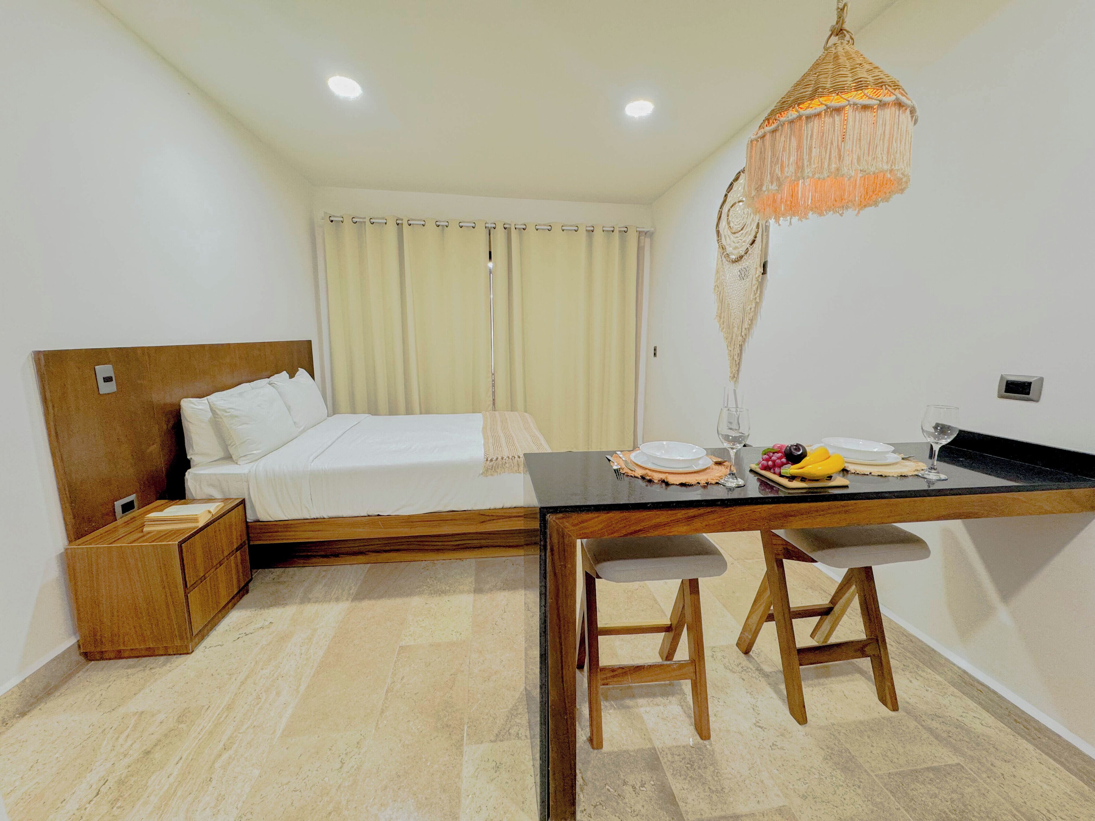 A hotel room with a bed, a desk with two chairs, a hanging light fixture, and a window with curtains.