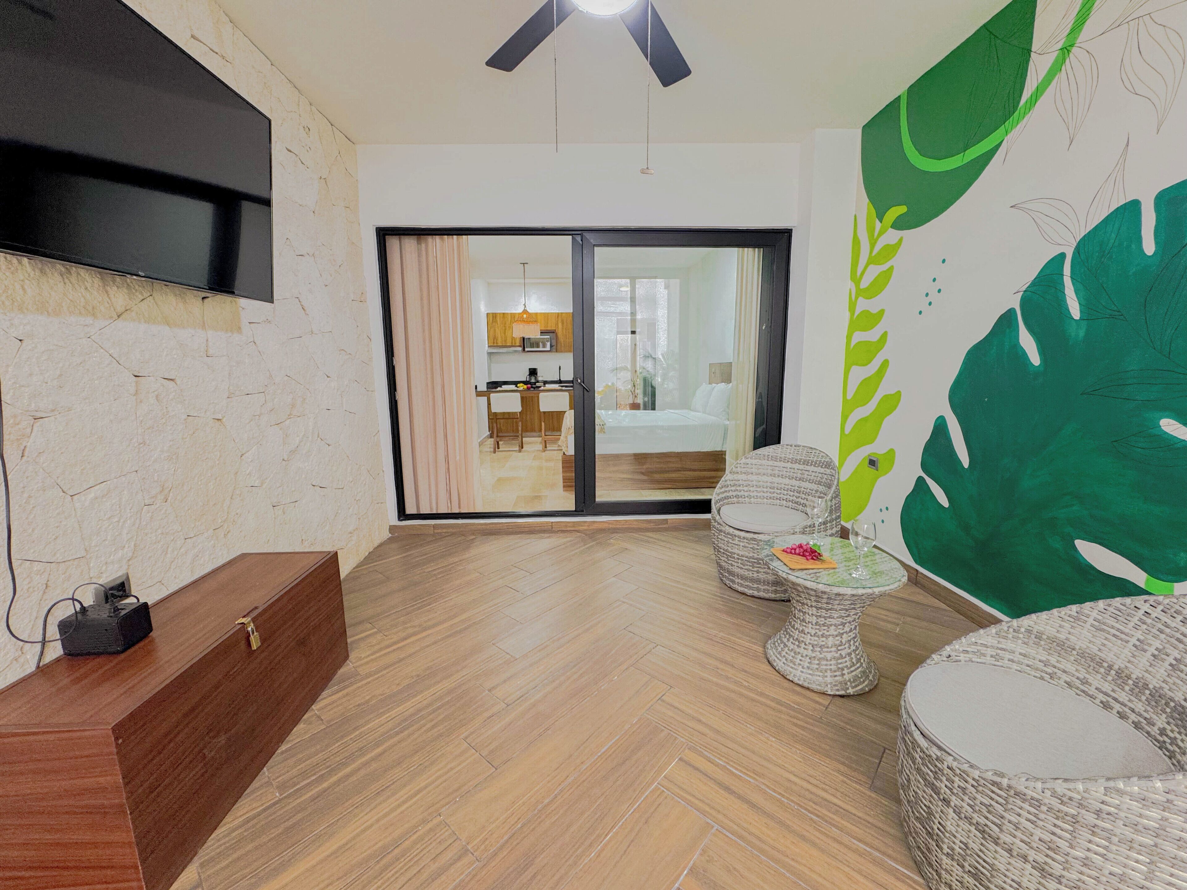 A hotel room with a wooden floor, a TV mounted on the wall, a bed with a sliding door, a dining area, and a large green leaf wall decoration.