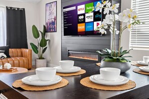 A modern living room with a fireplace, a television displaying streaming services, and a dining area with white bowls and plates. - Fun 5BR Theater, Games, and Spa! (North Las Vegas)