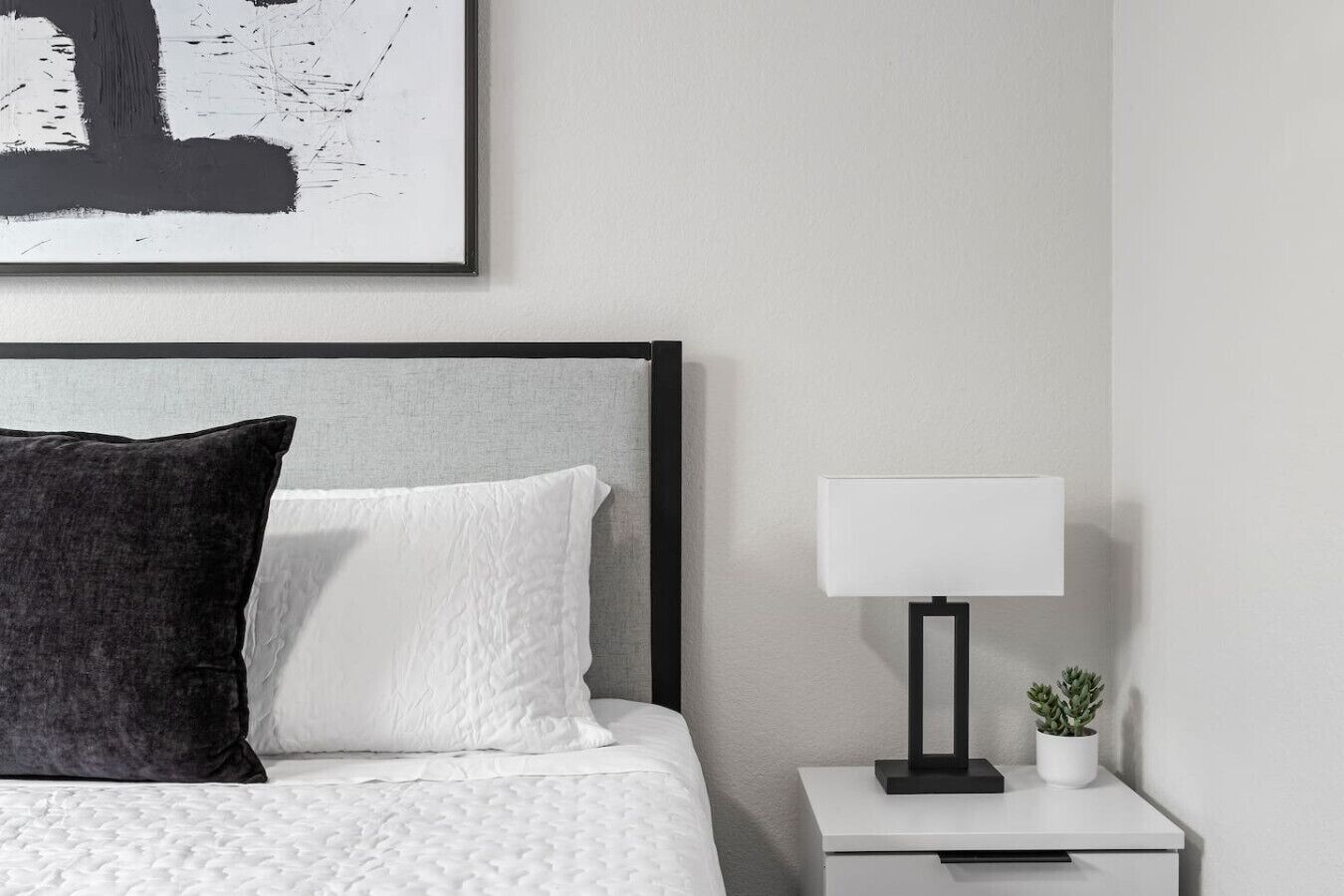 A neatly made bed with a grey headboard, a white pillow, and a black pillow. A white bedside table with a lamp and a small potted plant is beside the bed.
