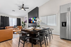 Een moderne woonkamer met een bank, salontafel en een eethoek met stoelen. Een flatscreen televisie is aan de muur bevestigd, en er is een koelkast in de achtergrond te zien.