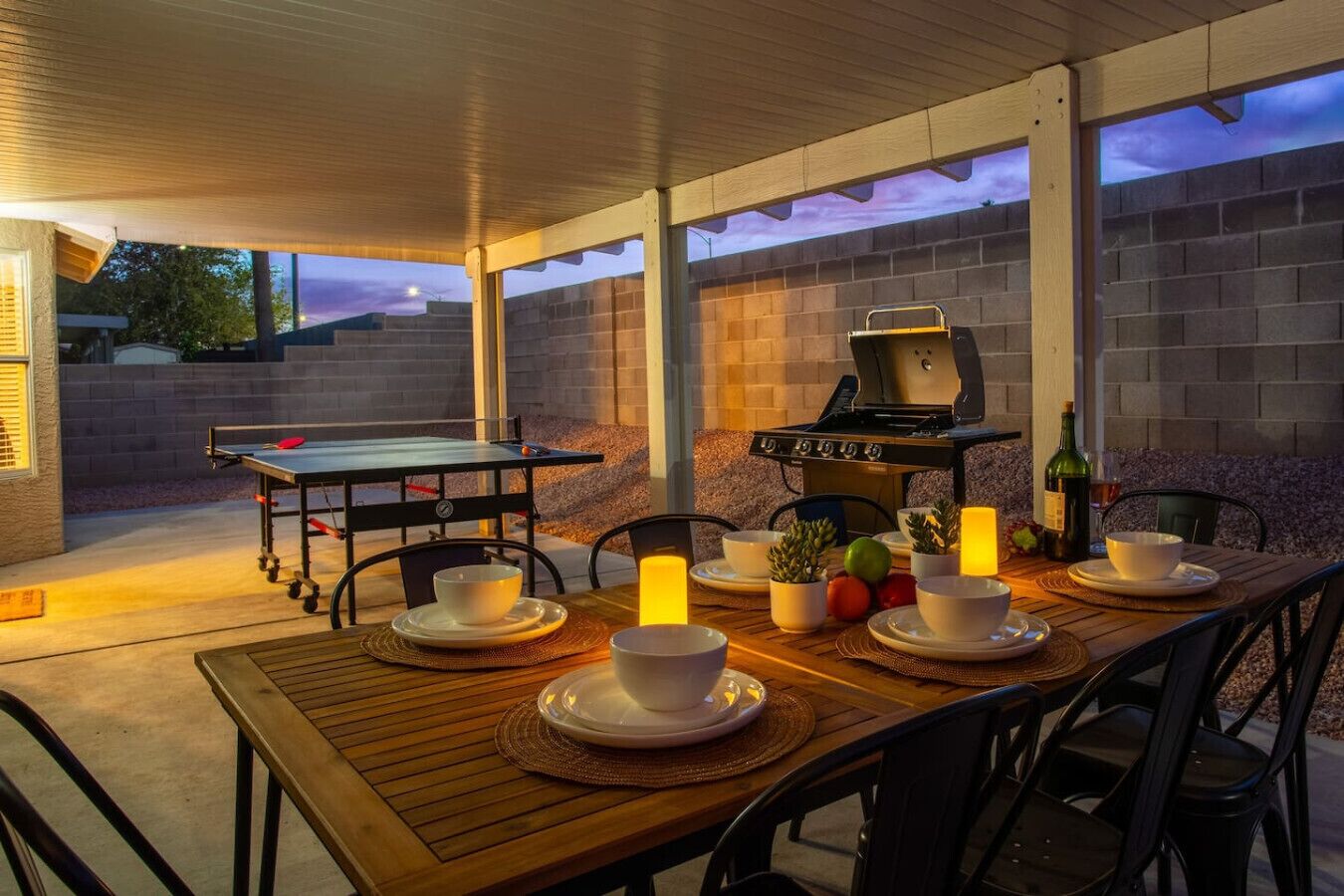 An outdoor patio with a wooden table set for dining, a grill, and a view of the backyard.
