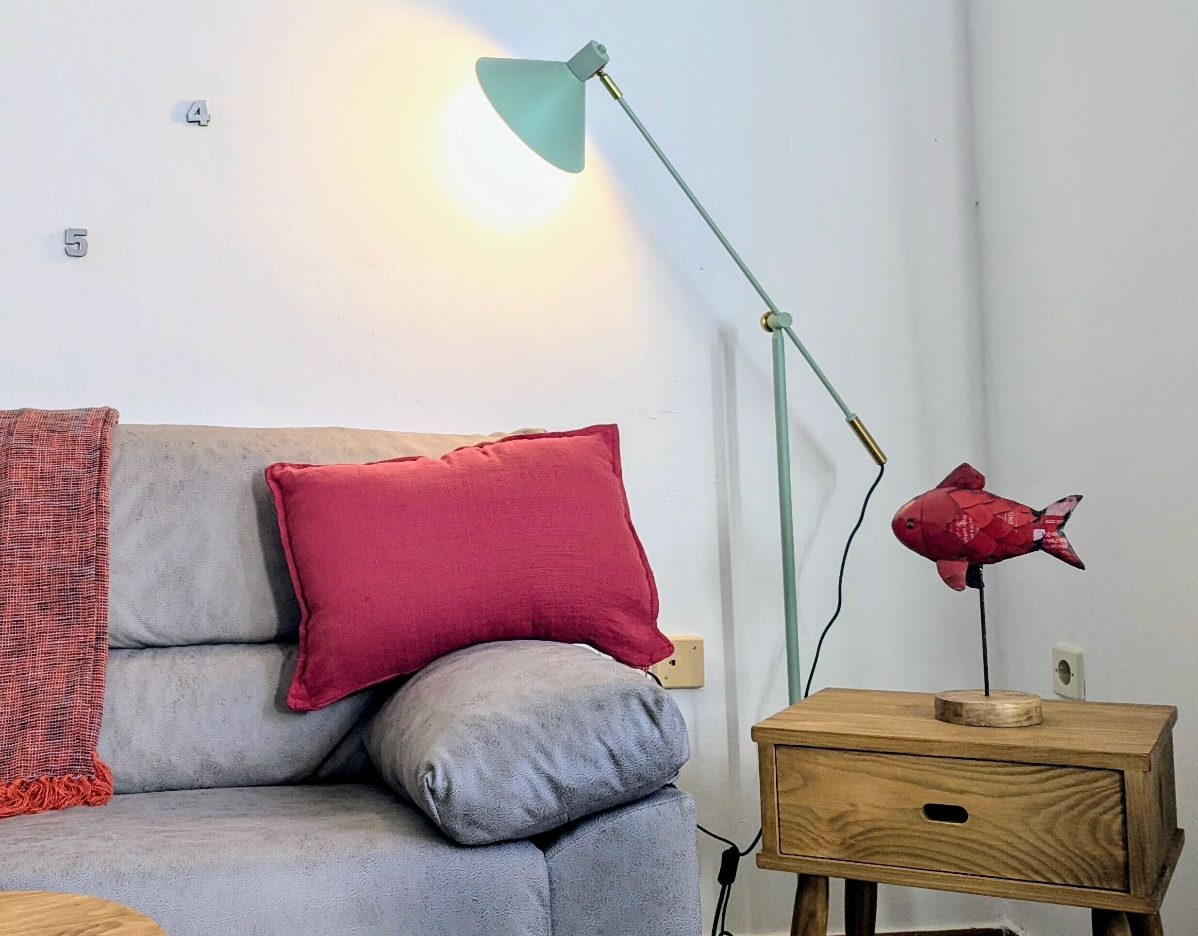 Ein Zimmer mit einem grauen Sofa, einem roten Kissen, einem Nachttisch aus Holz und einer grünen Stehlampe.