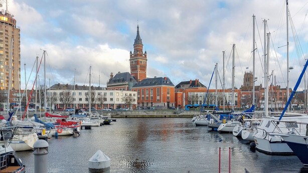 Een jachthaven met aangemeerde boten, een opvallende toren op de achtergrond en gebouwen langs de waterkant.