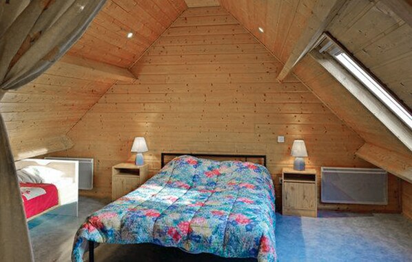 Un dormitorio con techo inclinado, paredes de madera, una cama con colcha colorida, dos mesitas de noche con lámparas y una ventana a la derecha.