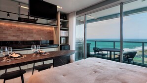 A modern hotel room with a large bed, a dining area with a table and chairs, a kitchen area with a TV, and a balcony with a view of the ocean. - RN Studio Premium 918 - Vista Mar  (Maceió)