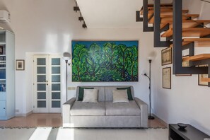 A modern living room with a beige sofa, a large abstract painting, a staircase, and a bookshelf. - Manara 29 - FromHometoRome (Roma)