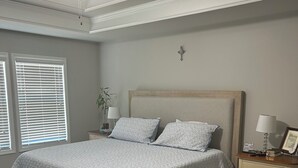 Une chambre à coucher comprenant un lit, une table de chevet, une lampe, un ventilateur de plafond et une fenêtre avec des stores.