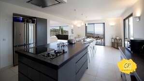 A modern kitchen with a central island, built-in appliances, and a view to the outdoors. - Villa d’exception 4 chambres, piscine, vue mer (Porto-Vecchio)