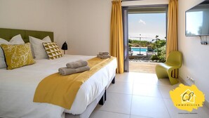 A hotel room with a bed, yellow curtains, a TV, and a view of a pool and greenery outside. - Villa d’exception 4 chambres, piscine, vue mer (Porto-Vecchio)