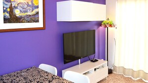 A room with a flat-screen TV mounted on the wall, a white cabinet below it, a patterned table, and a framed artwork above the TV.