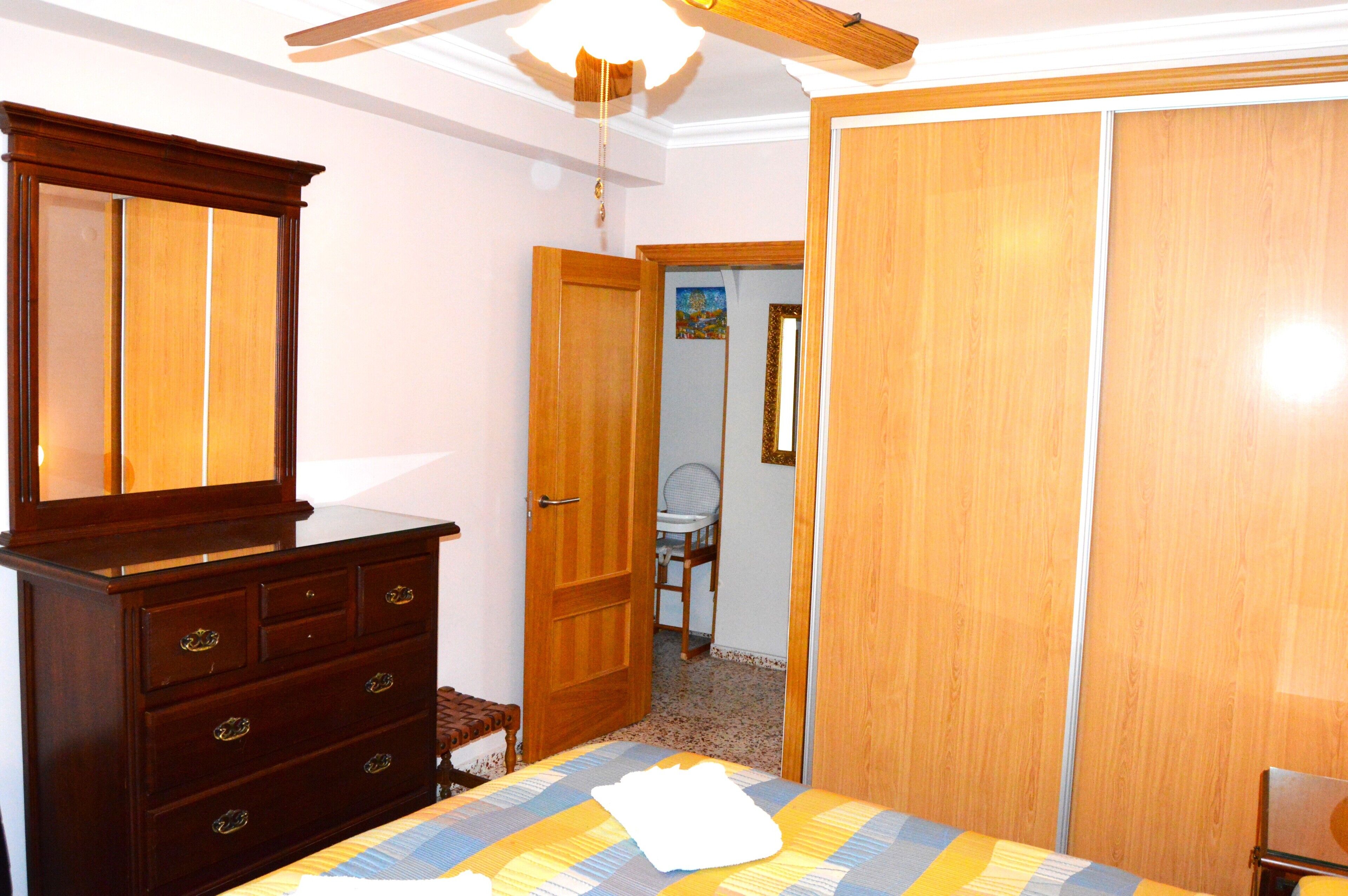 A bedroom with a wooden dresser, a bed with a patterned bedspread, a ceiling fan, and a wooden wardrobe.