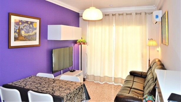 A room with purple walls, a dining table with chairs, a sofa, a TV, a lamp, and a window with curtains.