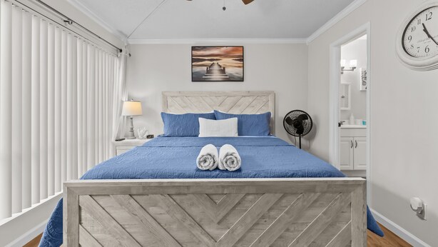 Une chambre à coucher avec un cadre de lit en bois, une literie bleue, une table de chevet avec une lampe, une horloge murale et un ventilateur de plafond.