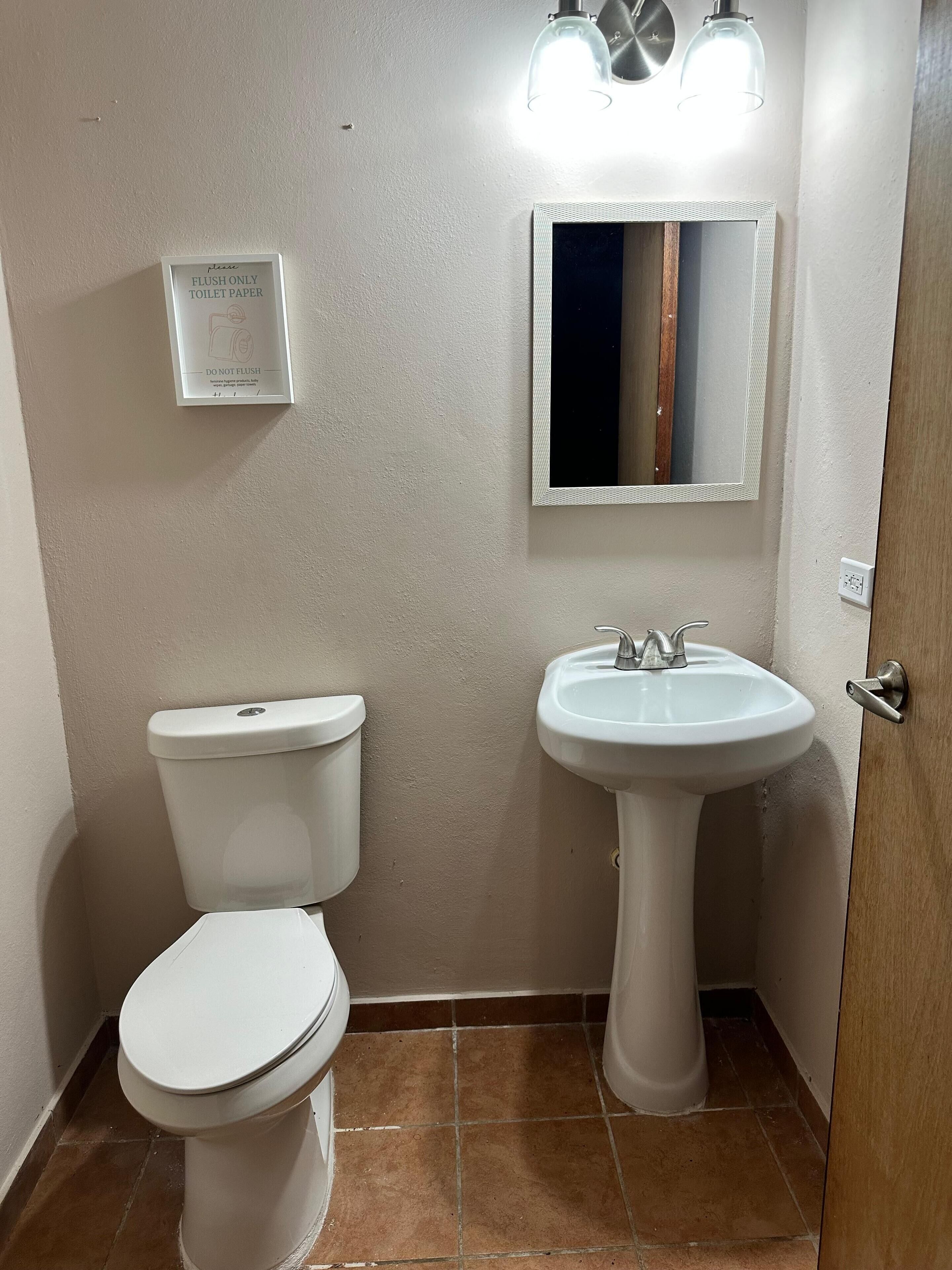 Et lille badeværelse med toilet, vask og spejl. Et skilt på væggen siger: "Skyl kun toiletpapir."