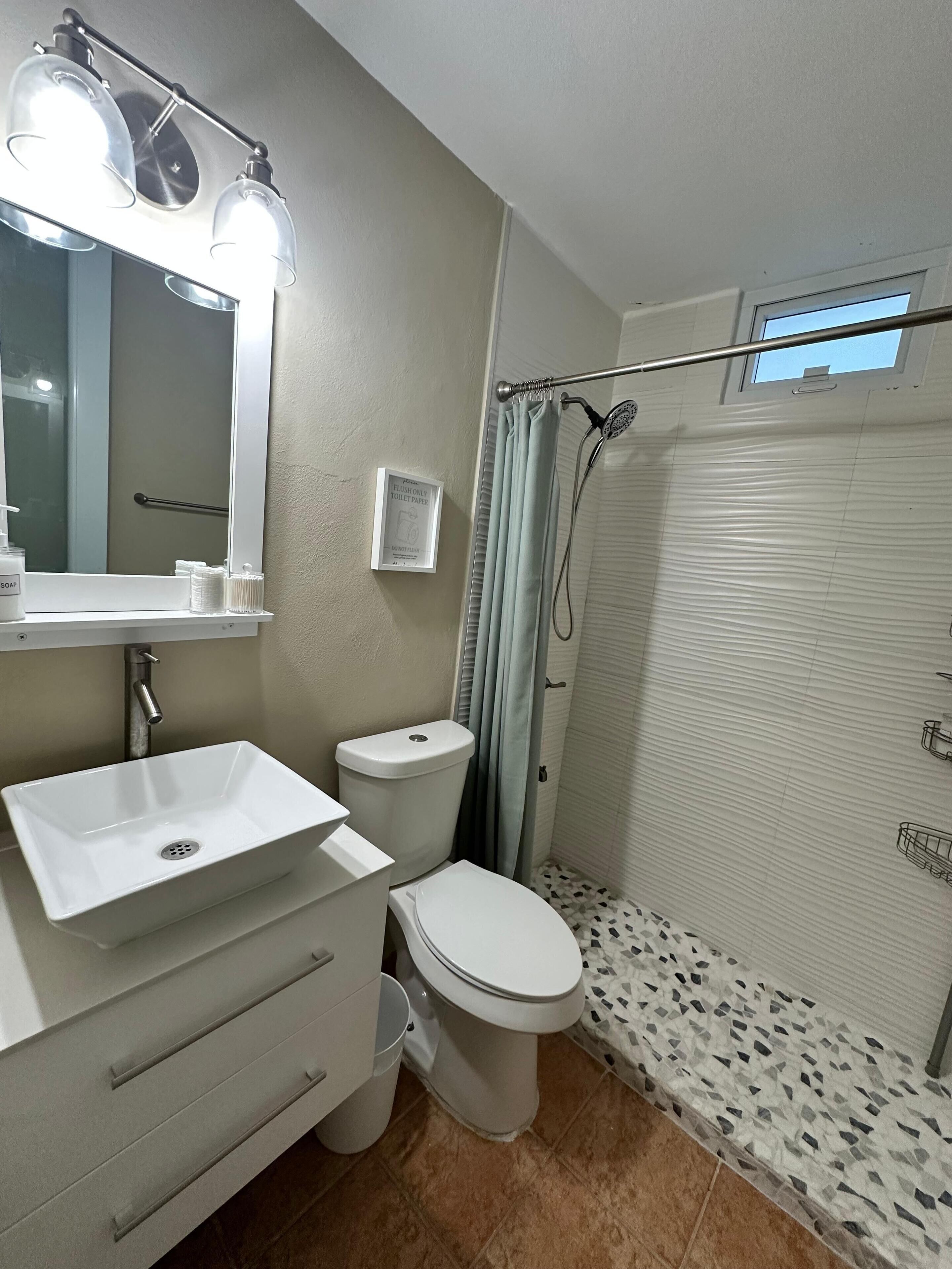 Un baño con lavamanos, inodoro y ducha blancos. Hay un espejo sobre el lavamanos y un letrero en la pared.
