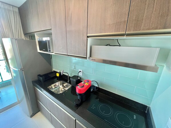 A modern kitchen with wooden cabinets, a stainless steel sink, a microwave, a red induction cooktop, and a window with curtains. - RN Studio Premium 1003 - Vista Mar (Maceió)