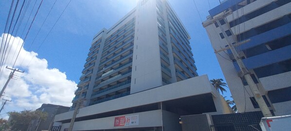 Un immeuble à plusieurs étages doté de balcons, une enseigne indiquant « RN Varada Gourmet Studio », et un ciel bleu clair parsemé de nuages.