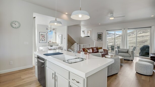 Une cuisine moderne avec des meubles blancs, un îlot central, des luminaires suspendus et une vue sur l’extérieur grâce à de grandes fenêtres.