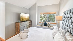 A bedroom with a bed, a TV on a wooden dresser, a window with a view of trees, and a ceiling fan.