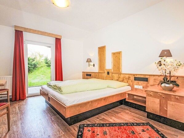 Une chambre avec un lit en bois, un petit bureau, une chaise, une table de chevet, une lampe et une vue sur la verdure à l’extérieur.