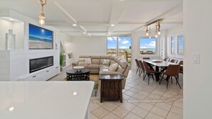 A spacious living area with a large sectional sofa, a dining table with chairs, a flat-screen TV mounted on the wall, and a large window offering a view of the beach. - Starboard Village 612 | Beachfront | Pensacola Beach | Pool | My Beach Getaways (Pensacola Beach)