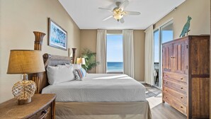 A bedroom with a bed, a wooden dresser, a ceiling fan, a nightstand, a lamp, and a window with a view of the ocean. - Spanish Key 307 | Beachfront | Steps from Flora Bama | Pools | My Beach Getaways (Pensacola)