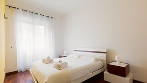 A neatly made bed with white linens and towels, a bedside table with a lamp, and a window with sheer curtains. - GuestHost - Milan Gambara Balcony Apartment (Milano)