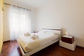 Een net opgemaakt bed met witte lakens en handdoeken, een nachtkastje met een lamp, en een raam met dunne gordijnen.