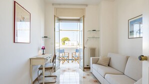 Une pièce avec un canapé blanc, un bureau avec une lampe, une chaise et une vue sur un espace extérieur aménagé avec une table et des chaises, accessible par une porte vitrée.