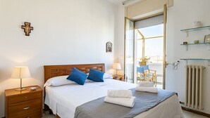 Un lit bien fait, orné d’oreillers bleus et blancs, une table de chevet en bois avec une lampe, une porte vitrée donnant sur l’extérieur et une petite étagère avec des objets décoratifs.